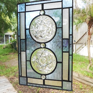 May include: A rectangular stained glass panel featuring a black lead frame, two circular clear glass inserts with starburst patterns, and textured light blue glass panes. The design includes vertical ribbed glass accents and three small clear glass cabochons.