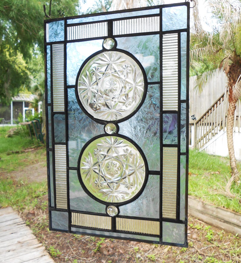 May include: A rectangular stained glass panel featuring a black lead frame, two circular clear glass inserts with starburst patterns, and textured light blue glass panes. The design includes vertical ribbed glass accents and three small clear glass cabochons.