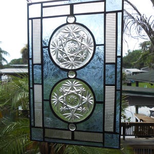 May include: A rectangular stained glass panel featuring two circular clear glass inserts with starburst patterns. The panel is framed with light blue textured glass, clear ribbed glass strips, and black metal lead lines, creating a geometric design.