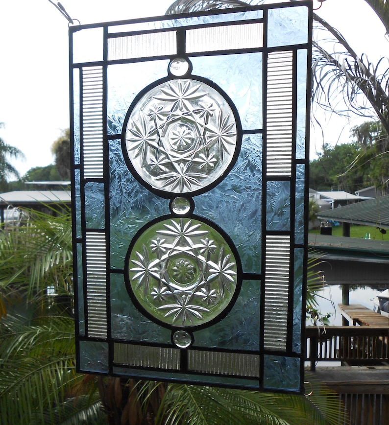 May include: A rectangular stained glass panel featuring two circular clear glass inserts with starburst patterns. The panel is framed with light blue textured glass, clear ribbed glass strips, and black metal lead lines, creating a geometric design.