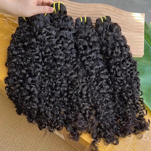 Può includere: Un fascio di extension per capelli ricci scuri. I capelli sono di un nero intenso e hanno una consistenza naturale e voluminosa. Le extension sono fissate con fasce gialle nella parte superiore.