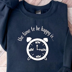 May include: A navy blue sweatshirt featuring the text "the time to be happy is..." and a white alarm clock design. The clock face displays "NOW" at the 12, 3, 6, and 9 o'clock positions. The sweatshirt has long sleeves.