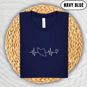 Puede incluir: Camiseta azul marino con un gr&aacute;fico blanco de un contorno de pa&iacute;s dentro de un dise&ntilde;o de latido de coraz&oacute;n. La camiseta se muestra sobre un mantel individual circular tejido. El texto "NAVY BLUE" est&aacute; en un &oacute;valo blanco.