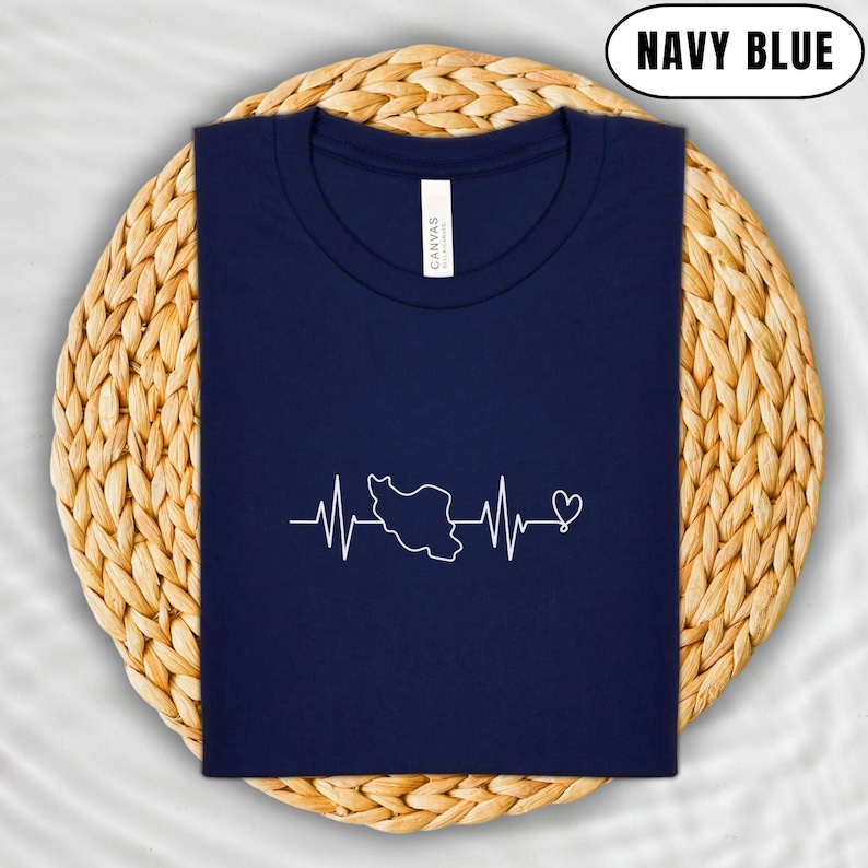 Puede incluir: Camiseta azul marino con un gr&aacute;fico blanco de un contorno de pa&iacute;s dentro de un dise&ntilde;o de latido de coraz&oacute;n. La camiseta se muestra sobre un mantel individual circular tejido. El texto "NAVY BLUE" est&aacute; en un &oacute;valo blanco.