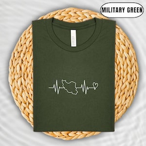 Puede incluir: Camiseta verde militar con un gr&aacute;fico blanco de una l&iacute;nea de latido del coraz&oacute;n con el contorno de un pa&iacute;s y un coraz&oacute;n. La camiseta est&aacute; doblada sobre un salvamanteles circular tejido.