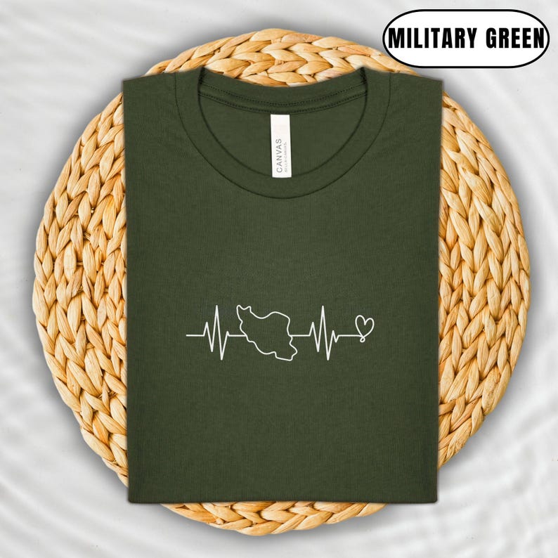 Puede incluir: Camiseta verde militar con un gr&aacute;fico blanco de una l&iacute;nea de latido del coraz&oacute;n con el contorno de un pa&iacute;s y un coraz&oacute;n. La camiseta est&aacute; doblada sobre un salvamanteles circular tejido.