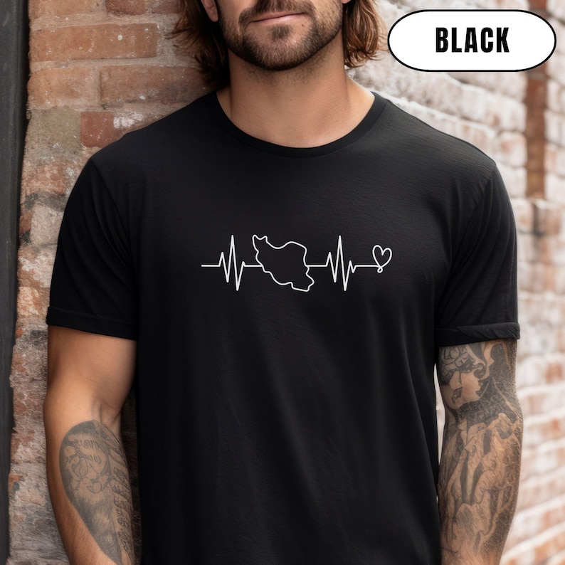 Puede incluir: Camiseta negra de cuello redondo con un gr&aacute;fico blanco de una l&iacute;nea de latidos, un mapa de Ir&aacute;n y un coraz&oacute;n. La palabra "BLACK" est&aacute; en un &oacute;valo blanco en la parte superior. La camiseta es de material suave.