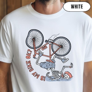 May include: White t-shirt featuring a cartoon skeleton and a bicycle with a bent wheel. The bicycle is red and grey. The text on the shirt reads "IS MY BIKE OK?"