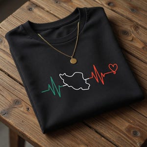 May include: A black crew neck t-shirt featuring a graphic print of a heartbeat line in green, white, and red. The line incorporates a white outline of the map of Iran in the center and ends with a red heart symbol. A gold chain necklace rests on top.