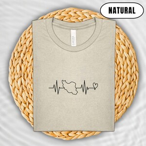 Puede incluir: Una camiseta beige con un gr&aacute;fico negro de una l&iacute;nea de latido card&iacute;aco con el contorno de un pa&iacute;s y un coraz&oacute;n. La palabra "NATURAL" est&aacute; en un &oacute;valo negro en la parte superior. La camiseta est&aacute; doblada sobre un salvamanteles circular tejido.