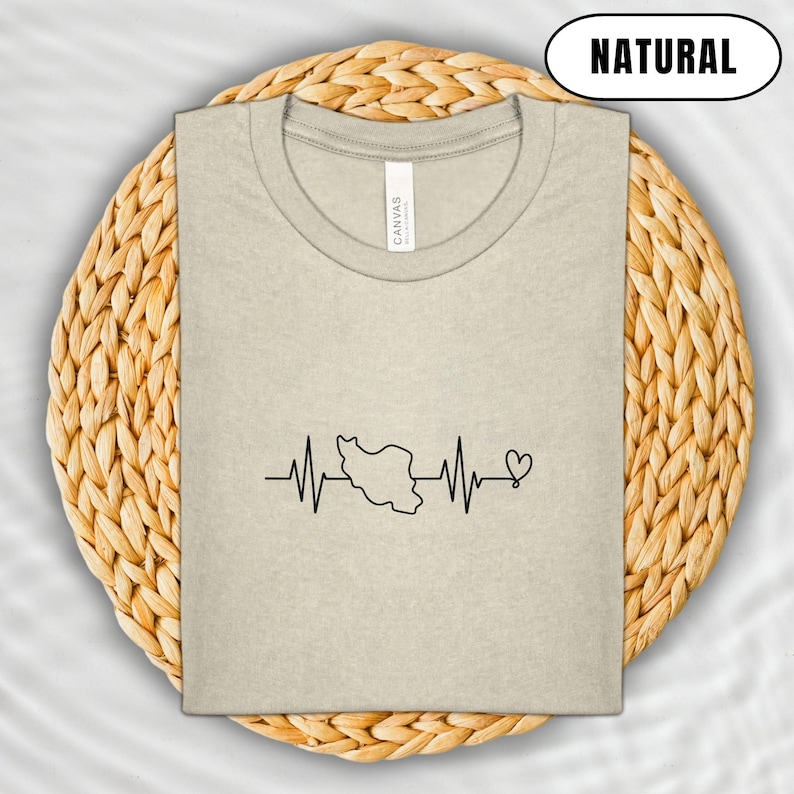 Puede incluir: Una camiseta beige con un gr&aacute;fico negro de una l&iacute;nea de latido card&iacute;aco con el contorno de un pa&iacute;s y un coraz&oacute;n. La palabra "NATURAL" est&aacute; en un &oacute;valo negro en la parte superior. La camiseta est&aacute; doblada sobre un salvamanteles circular tejido.