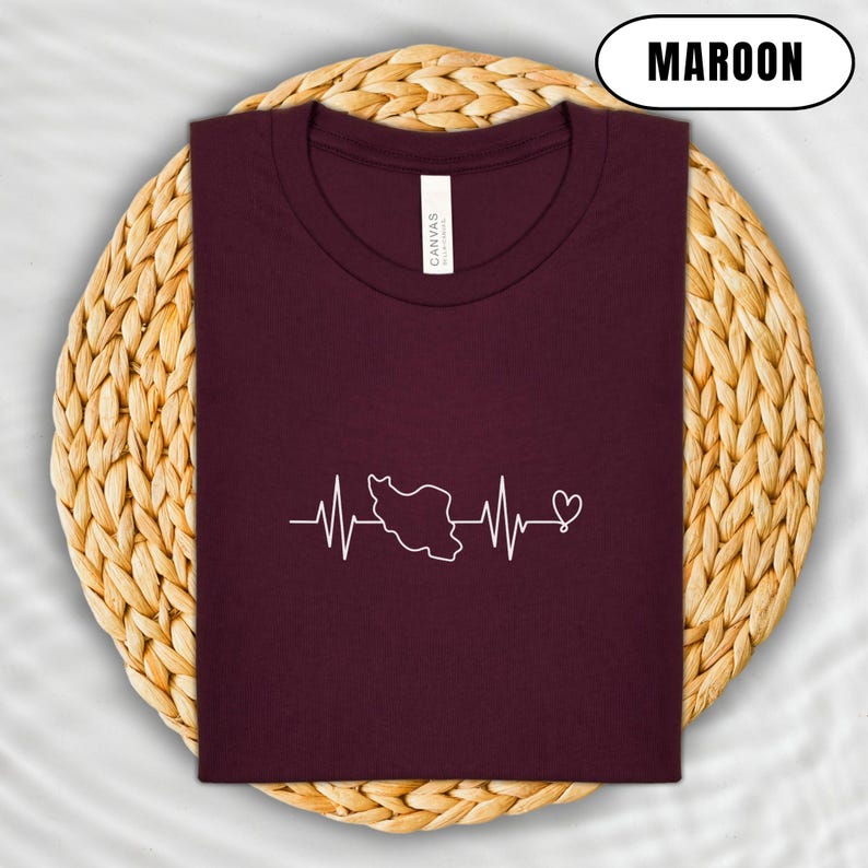 Puede incluir: Camiseta granate con un gr&aacute;fico blanco del contorno de un pa&iacute;s dentro de una l&iacute;nea de latido card&iacute;aco y un coraz&oacute;n. La camiseta est&aacute; doblada sobre un salvamanteles circular tejido.