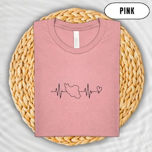 Puede incluir: Camiseta rosa claro con un dise&ntilde;o de l&iacute;nea de latido card&iacute;aco negro que presenta un mapa de Ir&aacute;n y un s&iacute;mbolo de coraz&oacute;n. La camiseta est&aacute; doblada sobre una estera circular tejida. La palabra "PINK" est&aacute; en un &oacute;valo blanco.