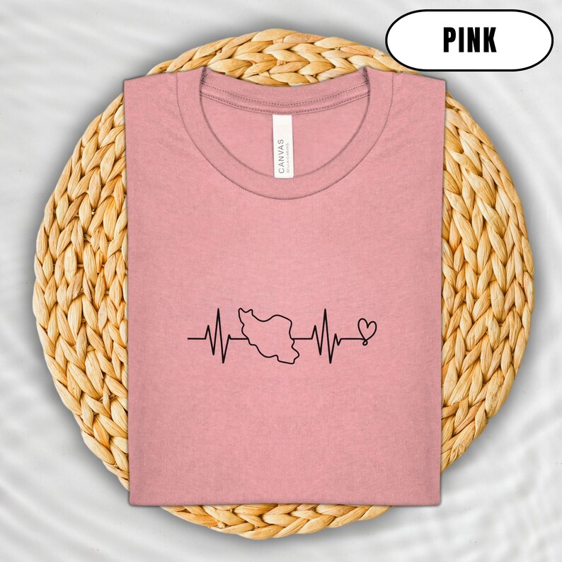 Puede incluir: Camiseta rosa claro con un dise&ntilde;o de l&iacute;nea de latido card&iacute;aco negro que presenta un mapa de Ir&aacute;n y un s&iacute;mbolo de coraz&oacute;n. La camiseta est&aacute; doblada sobre una estera circular tejida. La palabra "PINK" est&aacute; en un &oacute;valo blanco.