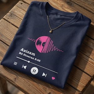 Peut inclure: Un t-shirt bleu marine avec un motif de disque vinyle rose et une interface de lecteur de musique imprimée. Le texte indique Azizam, Ed Sheeran 2:42, avec des commandes de lecture et un petit cœur rose. Une chaîne dorée repose sur le col.