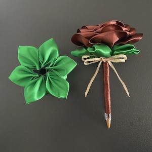 Puede incluir: Dos decoraciones florales hechas a mano con tela. Una es una flor verde de múltiples pétalos. La otra es una rosa marrón con hojas verdes, atada con una cinta beige y unida a un bolígrafo marrón.