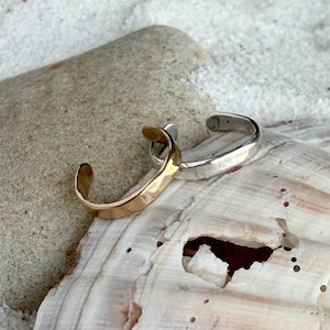 May include: Two open-ended cuff rings, one in a polished gold tone and one in a polished silver tone. Both rings feature a smooth, rounded band design with a gap at the center, allowing for adjustable sizing. The rings are metallic and reflective.
