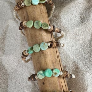 May include: Five bracelets made of wood and green and white beads. The bracelets are stacked on a piece of driftwood.