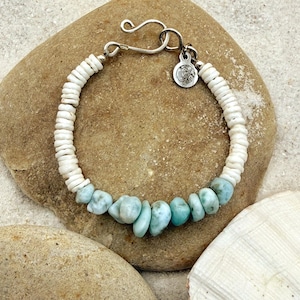 May include: A handmade bracelet featuring white shell beads and light blue larimar stones. The bracelet has a silver clasp and a small charm. The jewelry is displayed on a light brown rock, with a white seashell in the background.