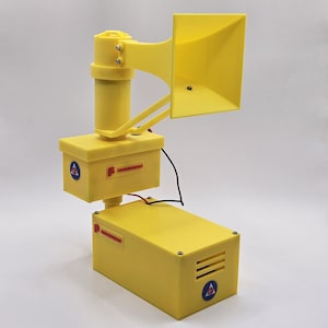 May include: A yellow electronic device with a horn-shaped speaker, a cylindrical component, and two rectangular boxes. The device has a blue and red logo on the boxes and a black wire. The device is likely a siren or alarm.
