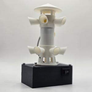 May include: A white, cylindrical device with multiple horn-shaped openings, mounted on a black rectangular base. The device has a switch and a power cord. The top has a circular, flat design. The overall design is modern and technical.