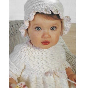 Puede incluir: Un bebé con un vestido y un gorro blancos de ganchillo. El vestido tiene un dobladillo y mangas con volantes, con un detalle de cinta rosa en la cintura. El gorro tiene un borde festoneado y un pequeño lazo.