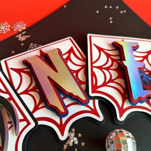 May include: A decorative banner featuring the letters N and E in a metallic iridescent finish, layered over white cardstock with a red spiderweb pattern. The letters are mounted on a black backing, creating a dimensional effect for a party display.