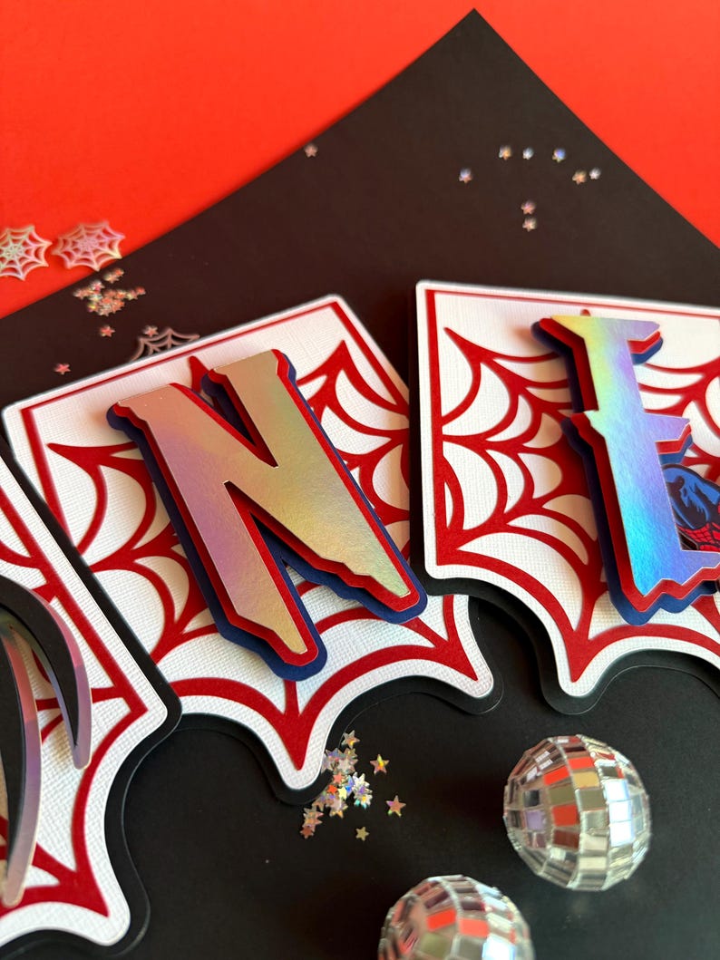 May include: A decorative banner featuring the letters N and E in a metallic iridescent finish, layered over white cardstock with a red spiderweb pattern. The letters are mounted on a black backing, creating a dimensional effect for a party display.
