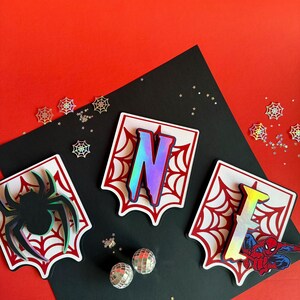 May include: Three white shield-shaped plaques with red spiderweb patterns. The left plaque features a black spider, the centre plaque displays the iridescent letter N, and the right plaque shows the iridescent letter E with a red and blue superhero.