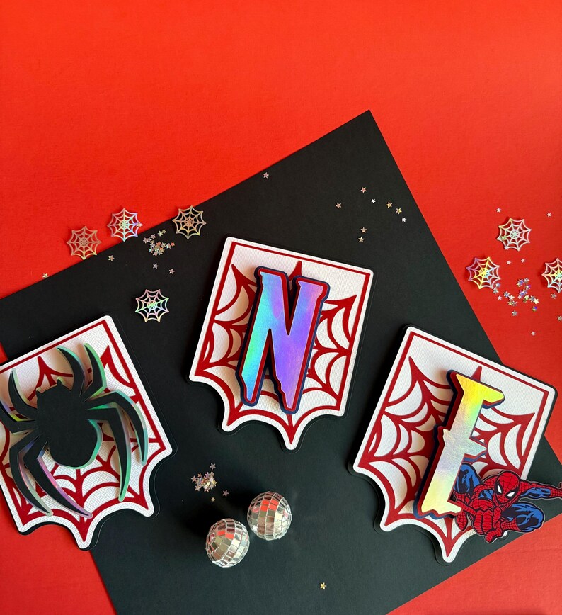 May include: Three white shield-shaped plaques with red spiderweb patterns. The left plaque features a black spider, the centre plaque displays the iridescent letter N, and the right plaque shows the iridescent letter E with a red and blue superhero.