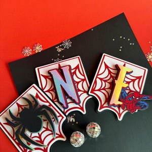 May include: Three decorative layered cutouts featuring a spider, the letter N, and the letter E. Each piece has a white background with a red spiderweb pattern. The letters have a holographic finish, and the spider is black with a holographic edge.
