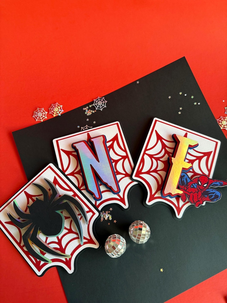 May include: Three decorative layered cutouts featuring a spider, the letter N, and the letter E. Each piece has a white background with a red spiderweb pattern. The letters have a holographic finish, and the spider is black with a holographic edge.