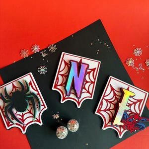 May include: Three white shield-shaped plaques with red spiderweb patterns. One features a black spider, one has the iridescent letter N, and one displays the iridescent letter I with a red and blue superhero character graphic.