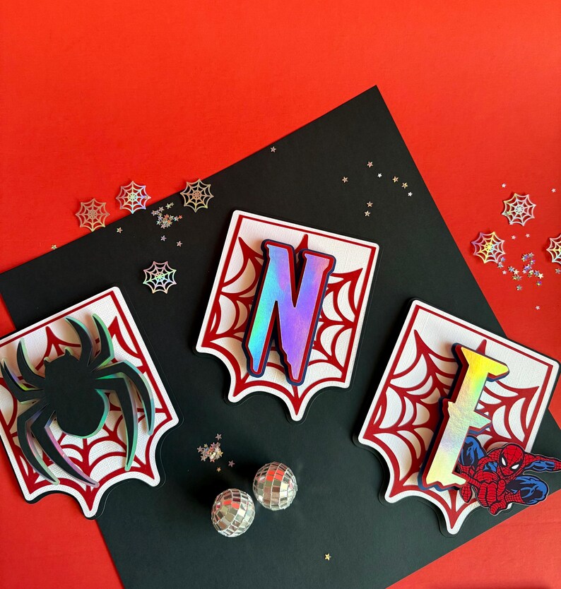 May include: Three white shield-shaped plaques with red spiderweb patterns. One features a black spider, one has the iridescent letter N, and one displays the iridescent letter I with a red and blue superhero character graphic.