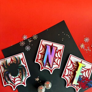 May include: Three white scalloped-edge plaques featuring red spiderweb patterns. The left plaque displays a black spider, the centre plaque features a large iridescent letter N, and the right plaque shows an iridescent letter E with a Spider-Man graphic.