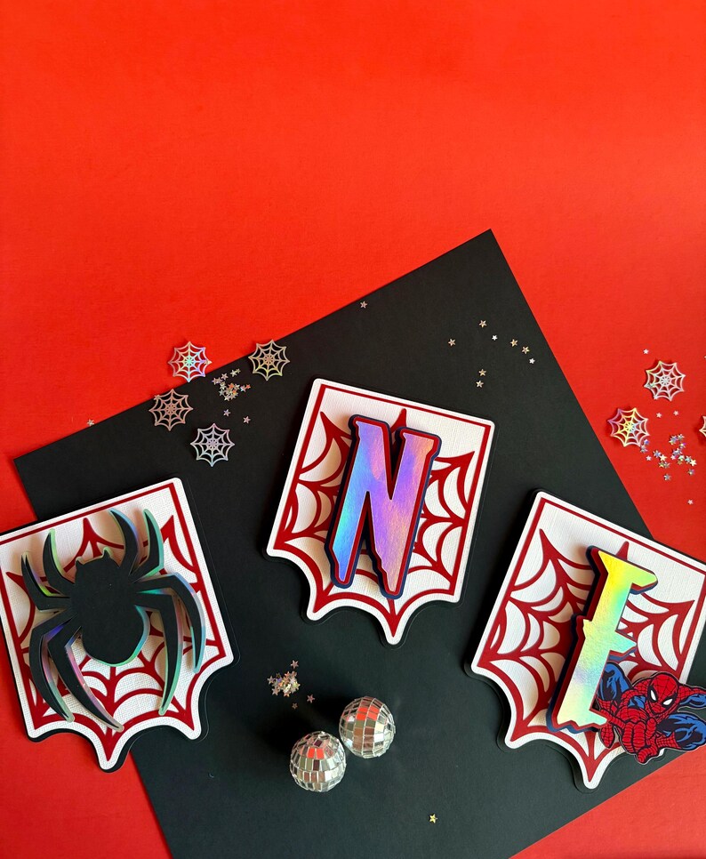 May include: Three white scalloped-edge plaques featuring red spiderweb patterns. The left plaque displays a black spider, the centre plaque features a large iridescent letter N, and the right plaque shows an iridescent letter E with a Spider-Man graphic.