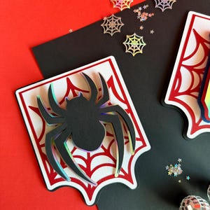 May include: A layered paper craft decoration featuring a black spider silhouette with iridescent edges centred on a white and red spiderweb design. The piece is accompanied by small holographic spiderweb and star-shaped confetti pieces.