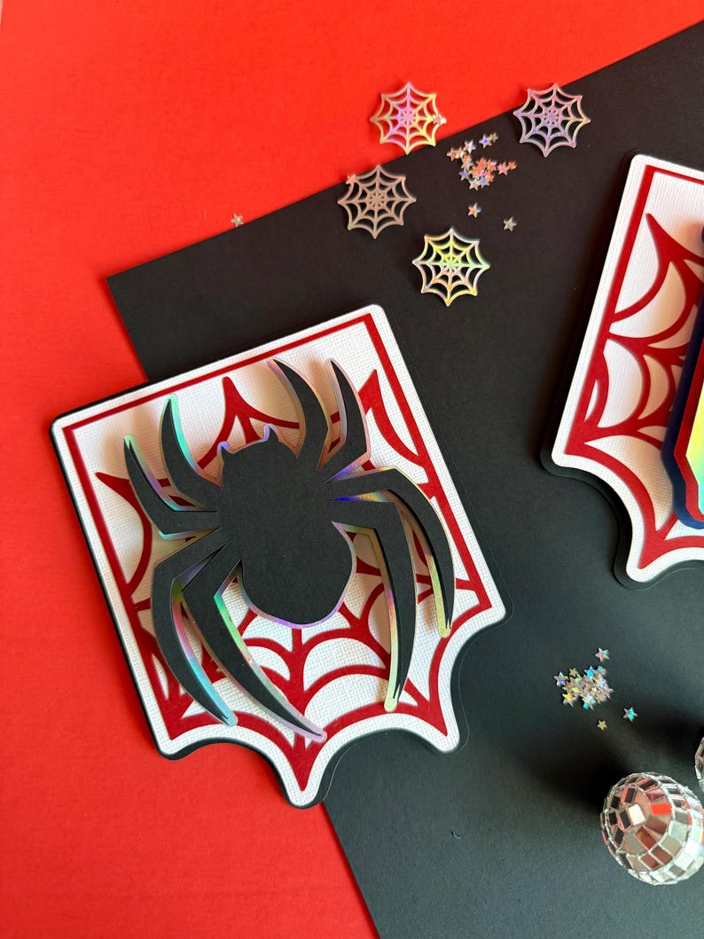 May include: A layered paper craft decoration featuring a black spider silhouette with iridescent edges centred on a white and red spiderweb design. The piece is accompanied by small holographic spiderweb and star-shaped confetti pieces.