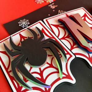 May include: A layered paper banner piece featuring a black spider cutout with iridescent edges, set against a white background with a red spiderweb pattern. The design includes a partial letter N with a gradient finish and small spiderweb accents.