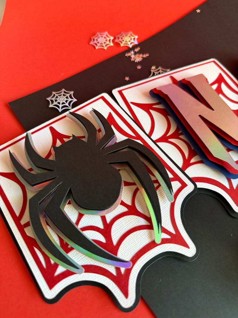 May include: A layered paper banner piece featuring a black spider cutout with iridescent edges, set against a white background with a red spiderweb pattern. The design includes a partial letter N with a gradient finish and small spiderweb accents.