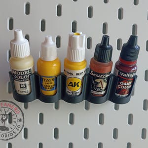 Puede incluir: Un soporte de plástico gris oscuro para botes de pintura con cinco botes de varios colores. Los botes están etiquetados con marcas como "Model Color" y "Game Color". El soporte está montado en una panel perforado blanco.