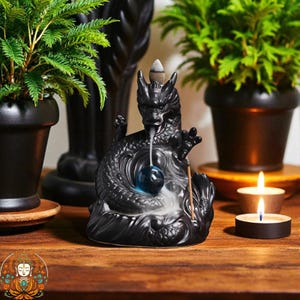 Puede incluir: Un quemador de incienso de dragón de cerámica negra con un orbe azul y un cono de incienso encendido. El dragón está rodeado de humo, con dos velas encendidas y plantas en macetas en el fondo.