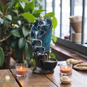 May include: A teal ceramic incense waterfall with cascading tiers, creating a visual of flowing smoke. Two lit candles in glass holders and small stones sit on a wooden surface, with lush green plants in the background. The incense burner is a decorative home decor item.