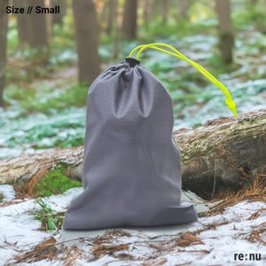 May include: A small gray drawstring bag with bright neon yellow cords. The bag is made of a smooth, solid-colored fabric and features the text Size // Small in the upper left corner and the brand name re:nu in the bottom right corner.