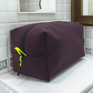May include: A rectangular dark purple toiletry bag featuring a black zipper closure and a bright neon yellow pull tab. The bag has a structured, boxy shape with clean lines and a smooth, solid-colored fabric exterior, providing a simple storage solution.