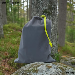 May include: A medium-sized gray drawstring bag featuring bright neon yellow cords. The bag has a smooth, solid-colored surface and is cinched at the top with the yellow drawstring. The text "Size // Medium" appears in the top left corner.