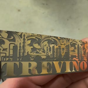 May include: A rectangular metal object with the name "TREVINO" in gold lettering. The object has an intricate design featuring industrial structures and decorative patterns in gold and dark gray.