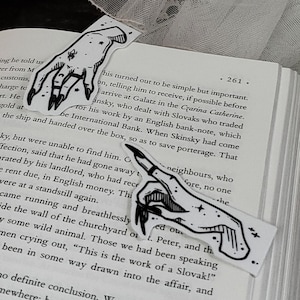 May include: Two black and white hand drawn bookmarks with a spooky, gothic aesthetic. The bookmarks are shaped like hands with long, sharp nails and stars. The bookmarks are placed on top of an open book with a candle in the background.