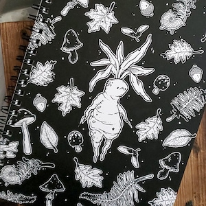 May include: Black and white spiral bound notebook with a whimsical illustration of a mushroom creature with leaves, mushrooms, and other botanical elements.