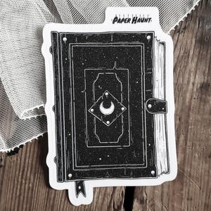 May include: Black and white illustration of a closed book with a crescent moon and stars design on the cover. The book is labeled "Paper Haunt."